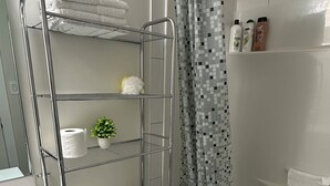 Une salle de bain avec des toilettes blanches, une douche avec un rideau à motifs, une étagère en métal avec des serviettes et une petite plante en pot.