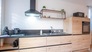 A modern kitchen with a black stove, a coffee maker, a microwave, and wooden cabinets. - Gite le palefrenier - 6 pers - 10 min Etretat (Gonneville-la-Mallet)