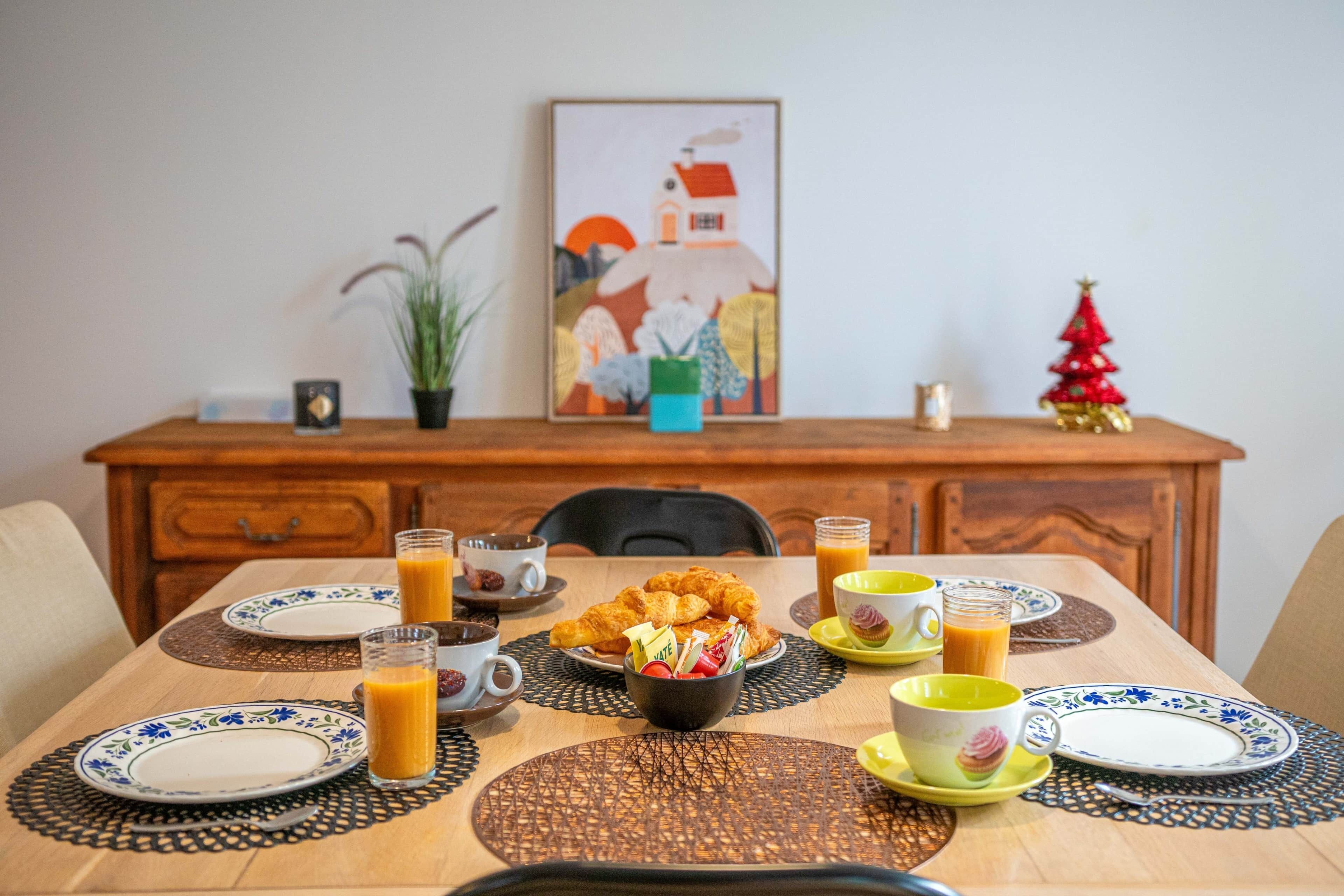 A dining table set with plates, cups, and a bowl of food, with a painting and a small Christmas tree on the wall behind.