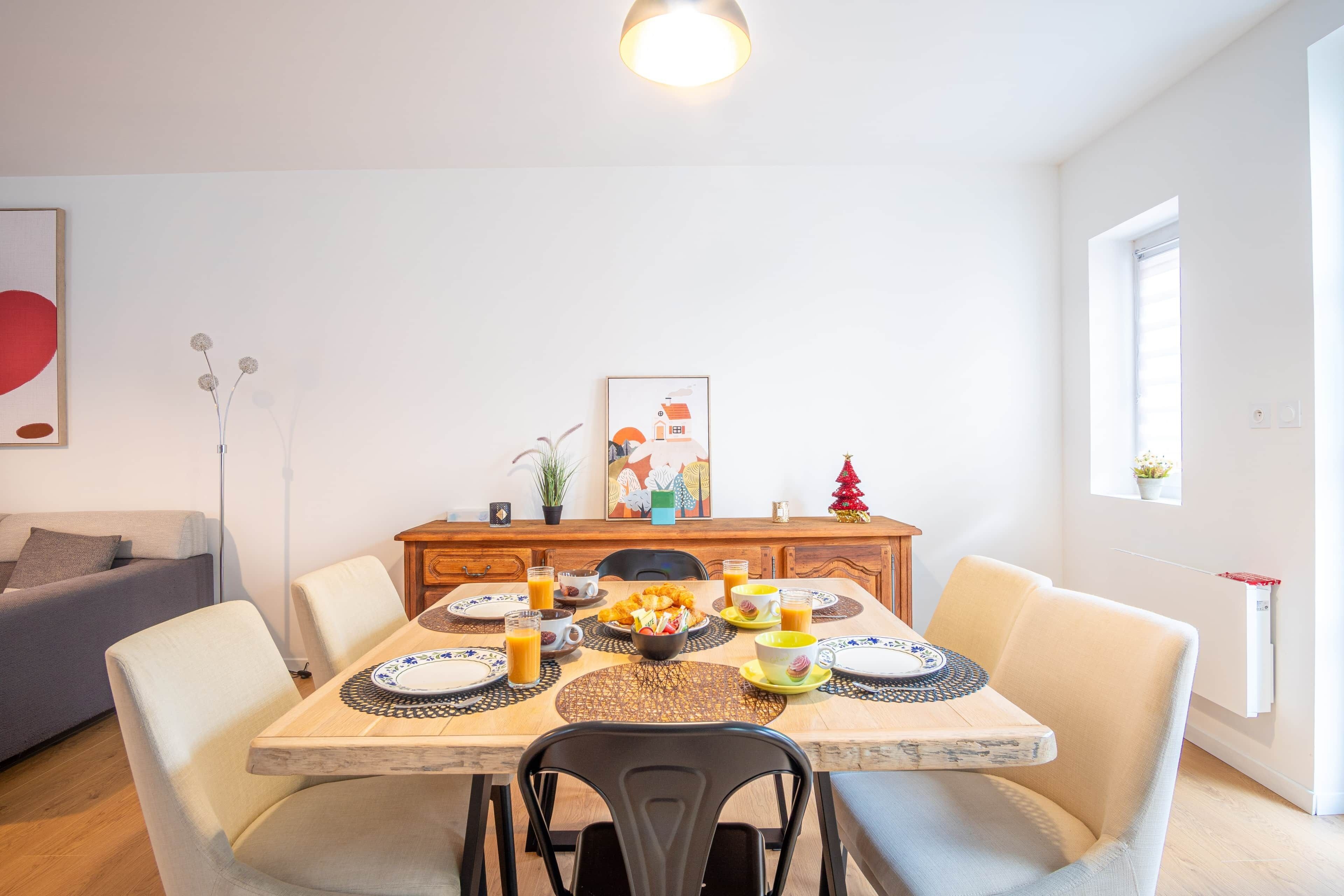 Ein Essbereich mit einem Holztisch, gedeckt für vier Personen, umgeben von Stühlen, mit einer Couch und einer kleinen Pflanze im Hintergrund.