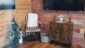 Un soggiorno accogliente con pavimento in legno, un'ampia TV a schermo piatto montata su un muro in mattoni, un albero di Natale e una poltrona confortevole.