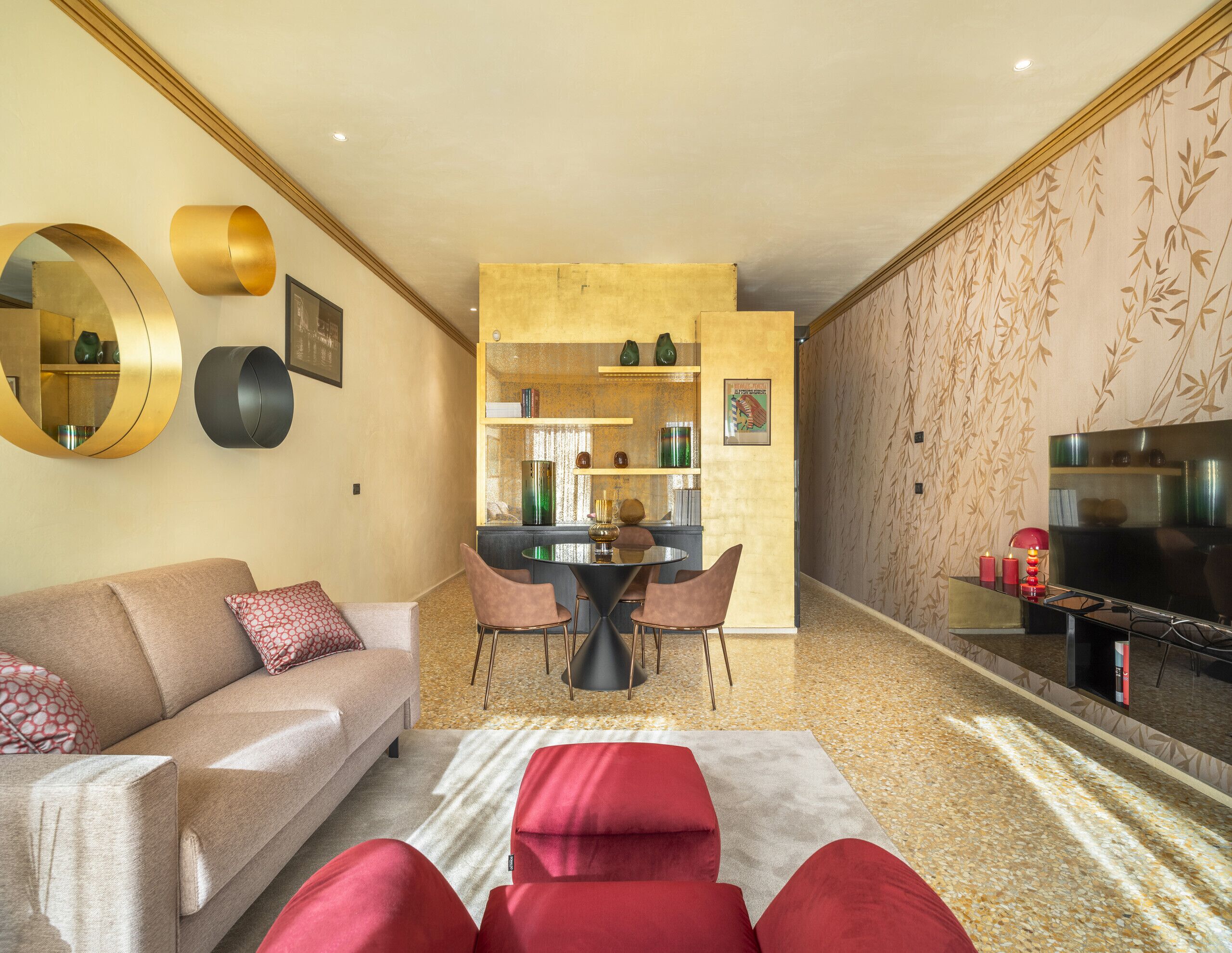 Un salon moderne avec un canapé, des coussins rouges, une table basse et un téléviseur. On y trouve également un coin repas avec une table et des chaises, ainsi qu’un coin cuisine avec des étagères et un réfrigérateur.