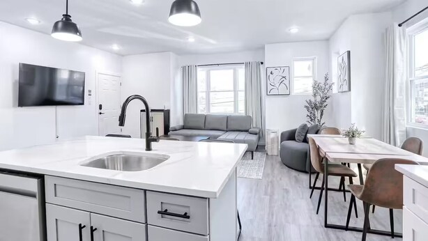 Et moderne køkken med en stor ø, et spiseområde med bord og stole, en grå sofa og et fjernsyn ophængt på væggen.