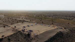 Luchtbeeld van een woonschap in een woestijn, met verschillende kleine gebouwen, waarvan sommige blauwe daken hebben, in een halve cirkel rond een centraal open gebied gerangschikt.