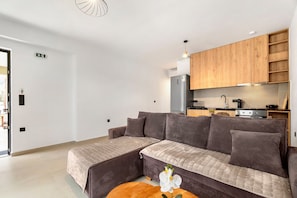 Una sala de estar moderna con un sofá, una mesita con una flor, y una zona de cocina con armarios de madera y electrodomésticos.