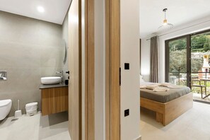 Une chambre moderne avec un lit en bois, une salle de bain dotée d’un lavabo blanc et une grande fenêtre donnant sur un paysage verdoyant.