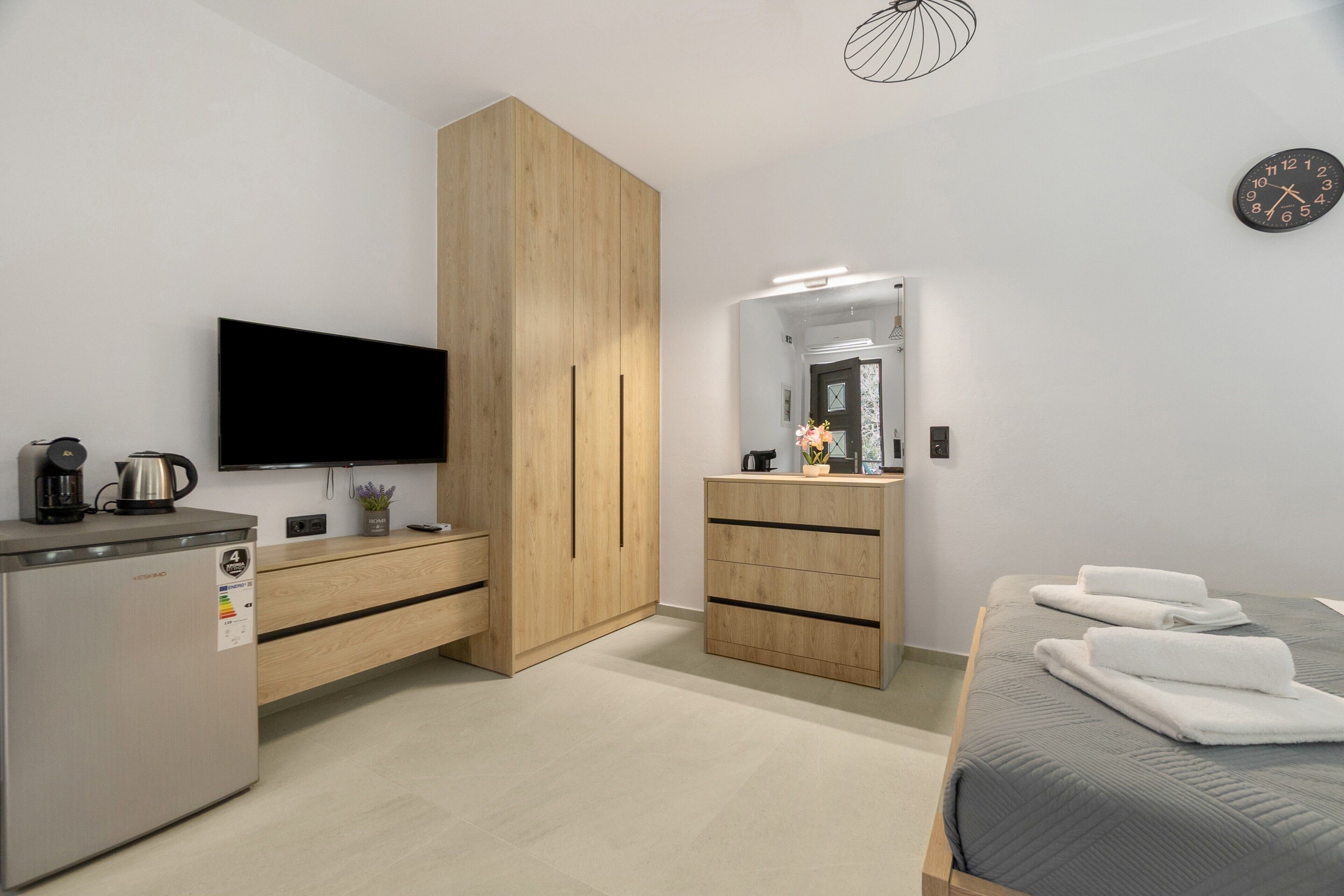 Een moderne slaapkamer met een flatscreen televisie, een houten garderobe, een klein kitchenette, een bed met handdoeken en een wandklok.