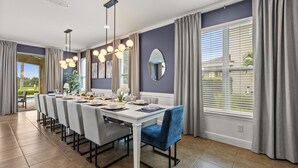 A dining area with a long table set for six, modern pendant lighting, and large windows with curtains. - Upstay Spacious 5BR Villa Private Pool and Hot Tub (Kissimmee)