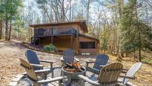 Eine Hütte mit einer Terrasse, einer Feuerstelle und Adirondack-Stühlen, vor einer bewaldeten Kulisse.