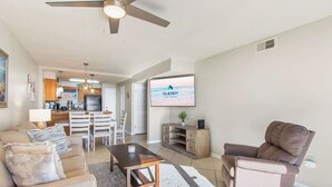 Un vaste salon doté d’un ventilateur de plafond, d’un téléviseur à écran plat, d’un canapé, d’une table basse et d’un coin repas avec une cuisine en arrière-plan.