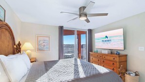 Une chambre à coucher comprenant un lit, un ventilateur de plafond, un téléviseur, une commode en bois et une fenêtre donnant sur un paysage agréable.