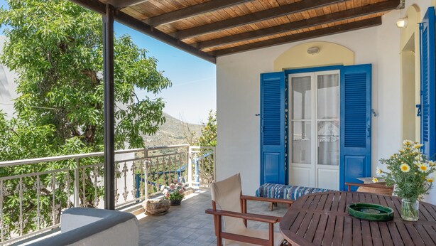 A wooden table with chairs on a balcony, a blue door, and a view of trees and hills. - Holiday Home 'Hippocrates' with Mountain View, Wi-Fi and Air Conditioning (Amorgos)