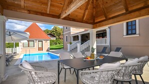 A covered outdoor area with a pool, wicker chairs, a table with fruits, and a view of a house with a red roof.