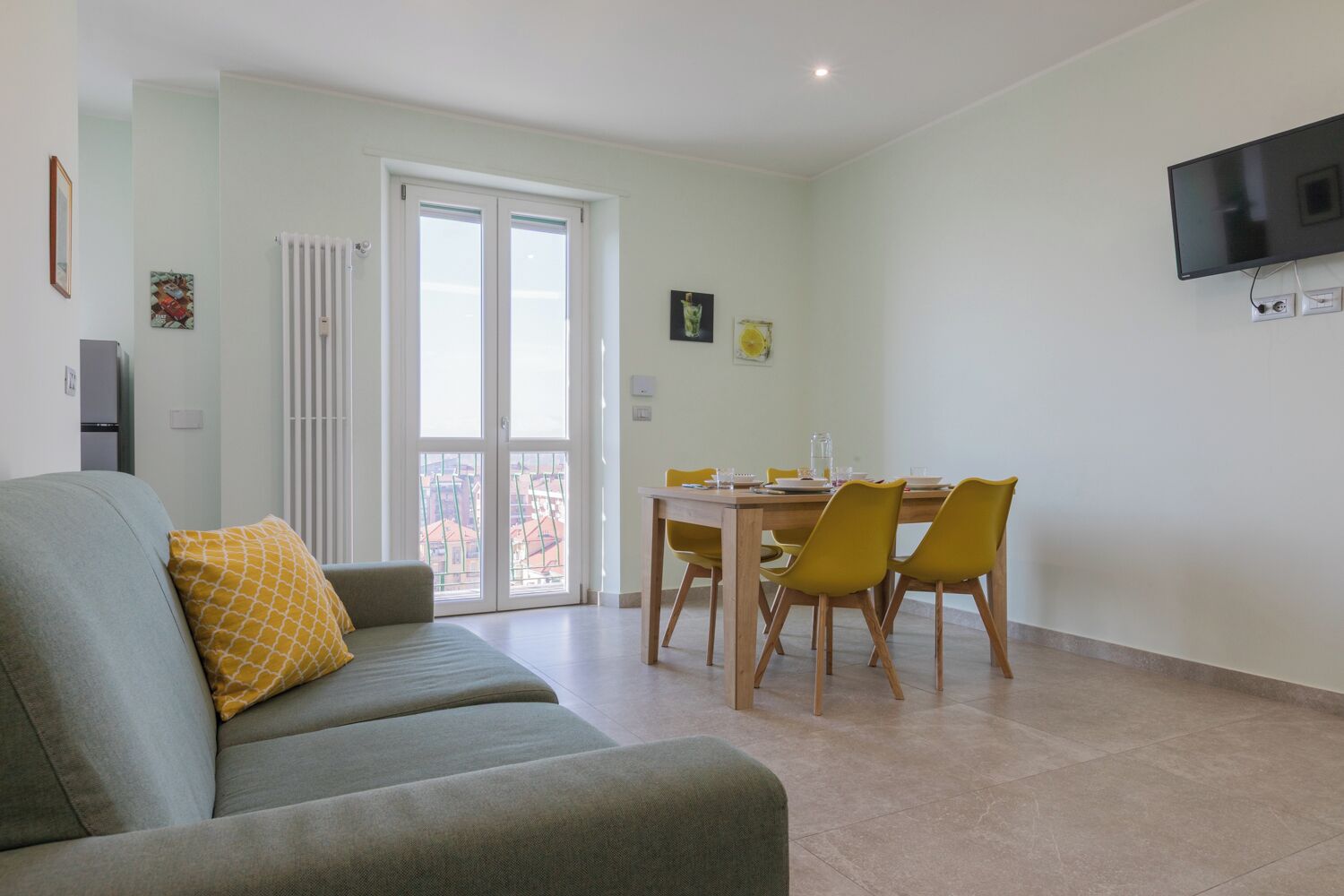Ein Wohnzimmer mit einer Couch, einem Esstisch mit Stühlen, einem an der Wand montierten Fernseher und einer Glasschiebetür, die zu einem Außenbereich führt.