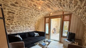 A cozy living room with a stone ceiling, a black sofa, a glass coffee table, and a TV on a wooden cabinet. - Mas Héritage (Rosières)