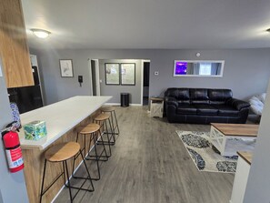 Ett modernt kök med en vit bänkskiva och barstolar, en svart lädersoffa och en mönstrad matta.