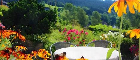 Un jardín con una mesa y sillas, rodeado de diversas flores y una vista de una colina boscosa.