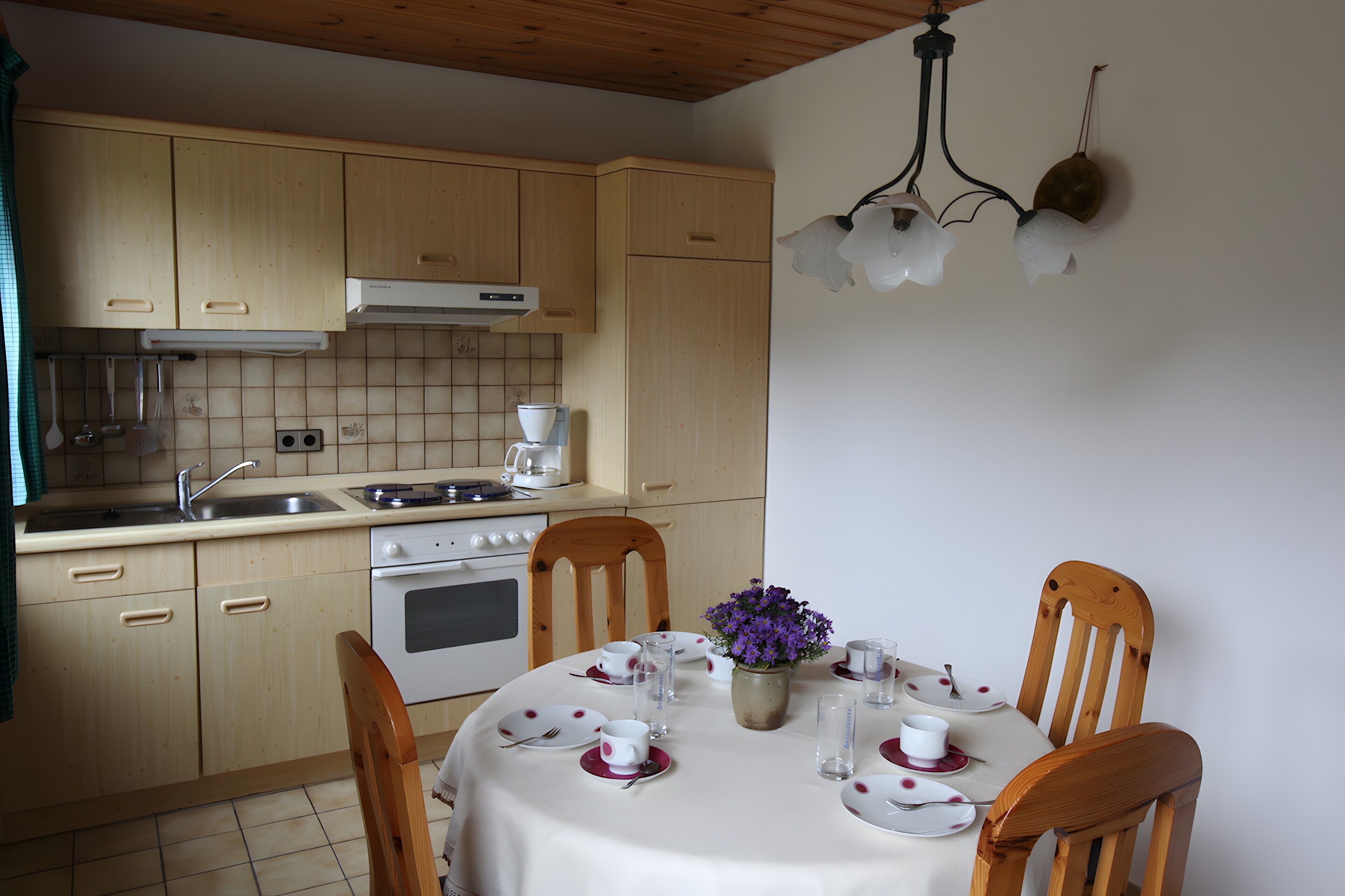 Une cuisine avec des meubles de rangement en bois, une cuisinière et une table à manger dressée pour deux personnes, avec des assiettes, des tasses et un vase de fleurs.