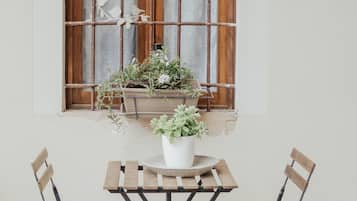 A window with wooden bars and a curtain, two wooden chairs, a small round table, and potted plants on the windowsill.