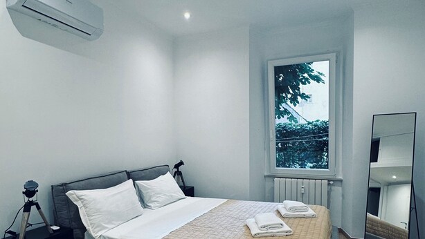 A modern bedroom with a bed, a nightstand, a chair, a window, and a mirror.