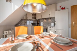 A dining area with a table set for four, a kitchen in the background, and a yellow pendant light above. - Ferienwohnung in Westerdeichstrich (Westerdeichstrich)