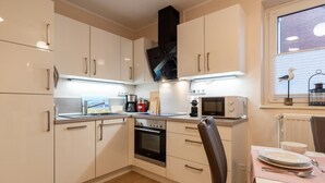 A compact kitchen with white cabinets, a built-in oven, a microwave, and a window with blinds. - Ferienwohnung in Büsum (Büsum)