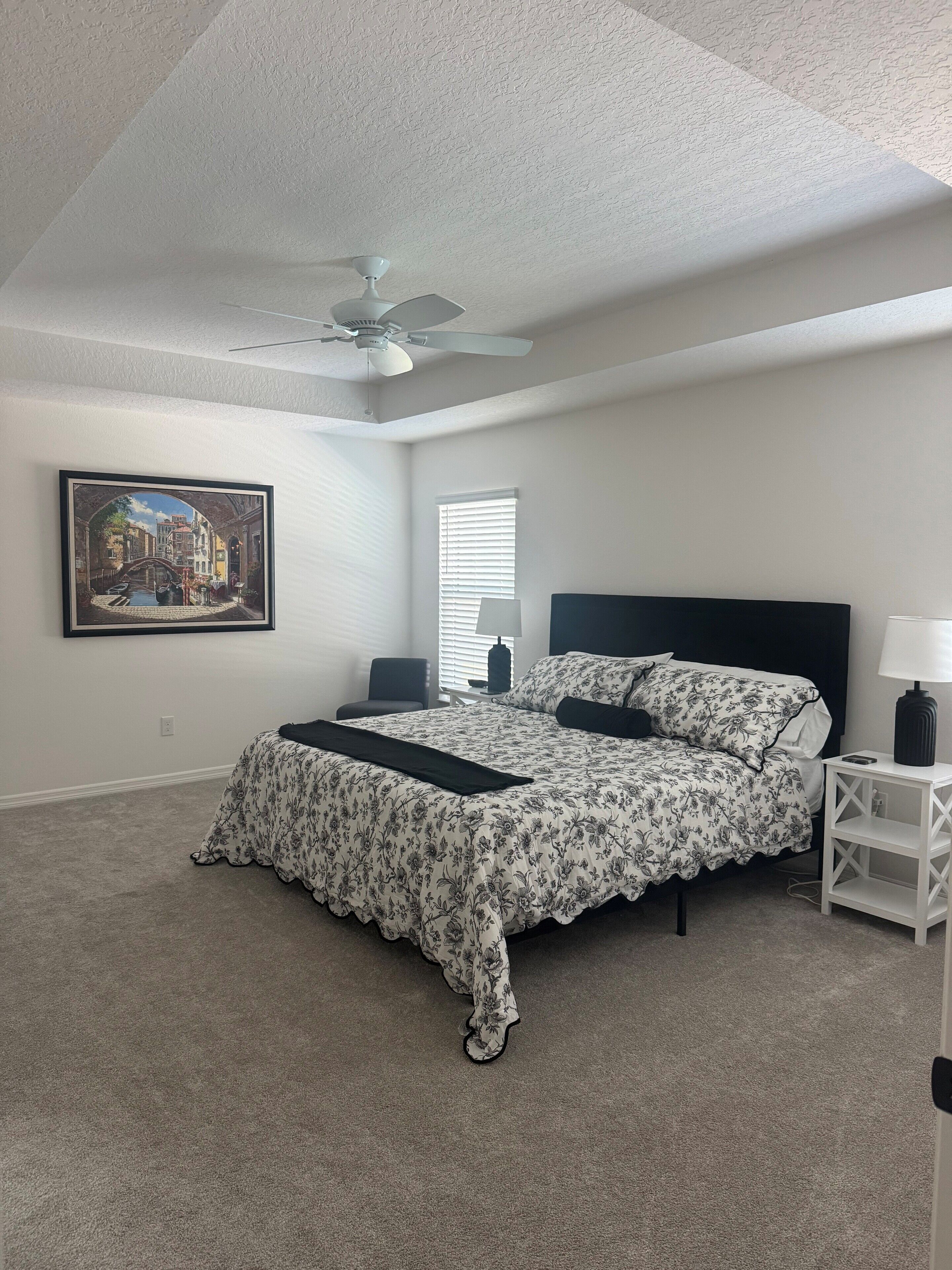 A bedroom with a bed, a ceiling fan, a nightstand, a lamp, a framed picture on the wall, and a window with blinds.