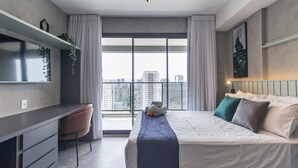 Une chambre moderne avec un lit, un bureau et une chaise. Une grande fenêtre, ornée de rideaux, offre une vue sur la ville.