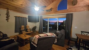 Un accogliente soggiorno con una ventola a soffitto, una testa di cervo appesa al muro, un televisore e vista sul tramonto attraverso la porta vetrata.