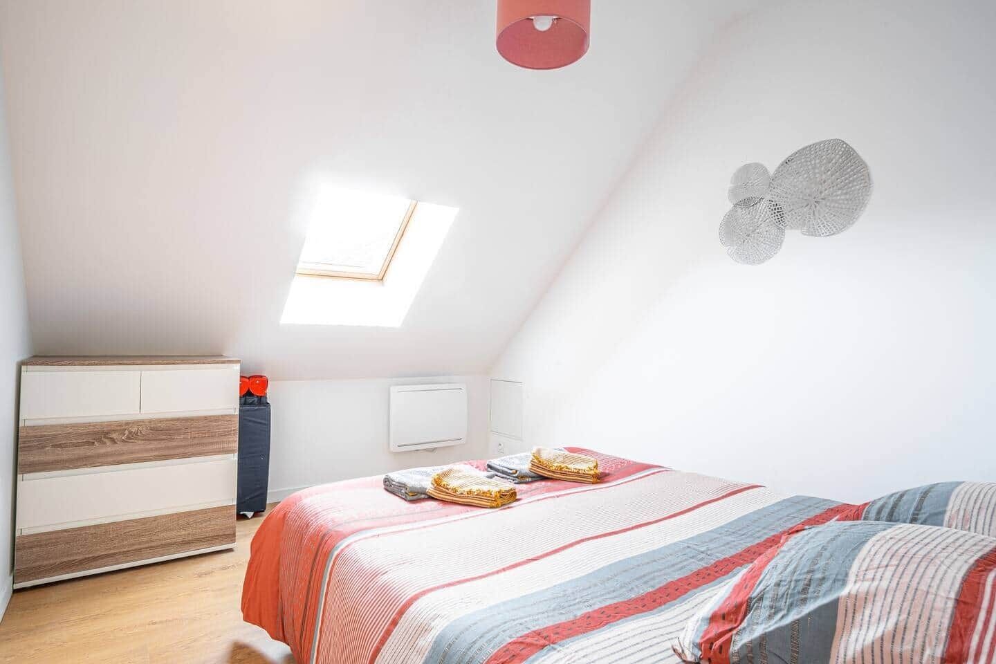 A bedroom with a sloped ceiling, a bed with striped bedding, a bedside table, a radiator, and a window.