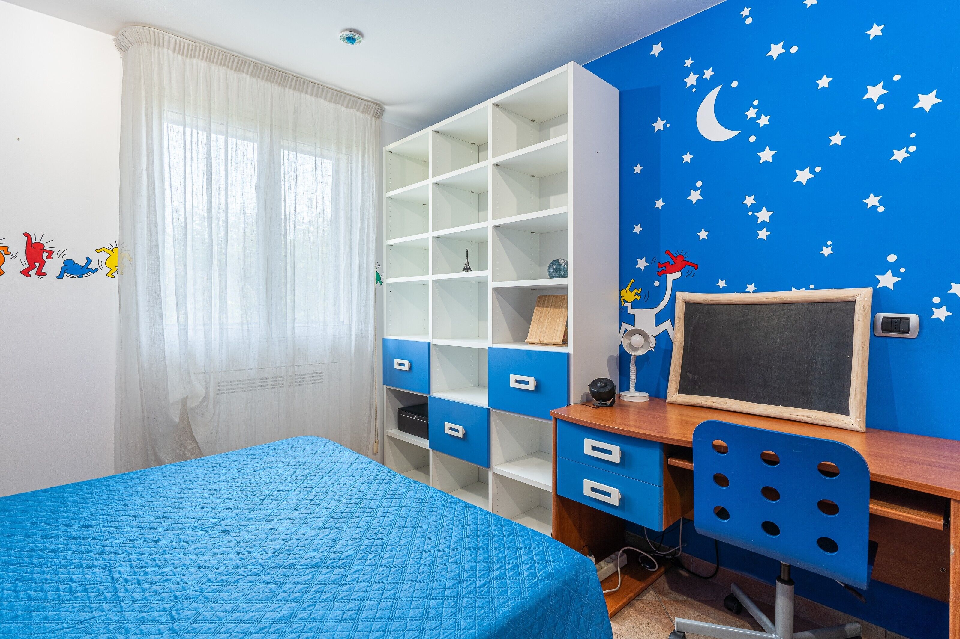 A bedroom with a bed, a desk with a chair, a bookshelf, and a wall with star and moon decals.