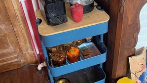 Un carrello da cucina blu con ripiano superiore in legno che contiene una macchina da caffè, un bollitore e una tazza rossa. Nella parte inferiore, diversi oggetti da cucina e un cestino giallo.