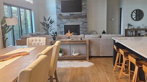 Ein modernes Wohnzimmer mit Kamin aus Stein, einem großen an der Wand montierten Fernseher und einem Essbereich mit Holzsesseln und Tisch.