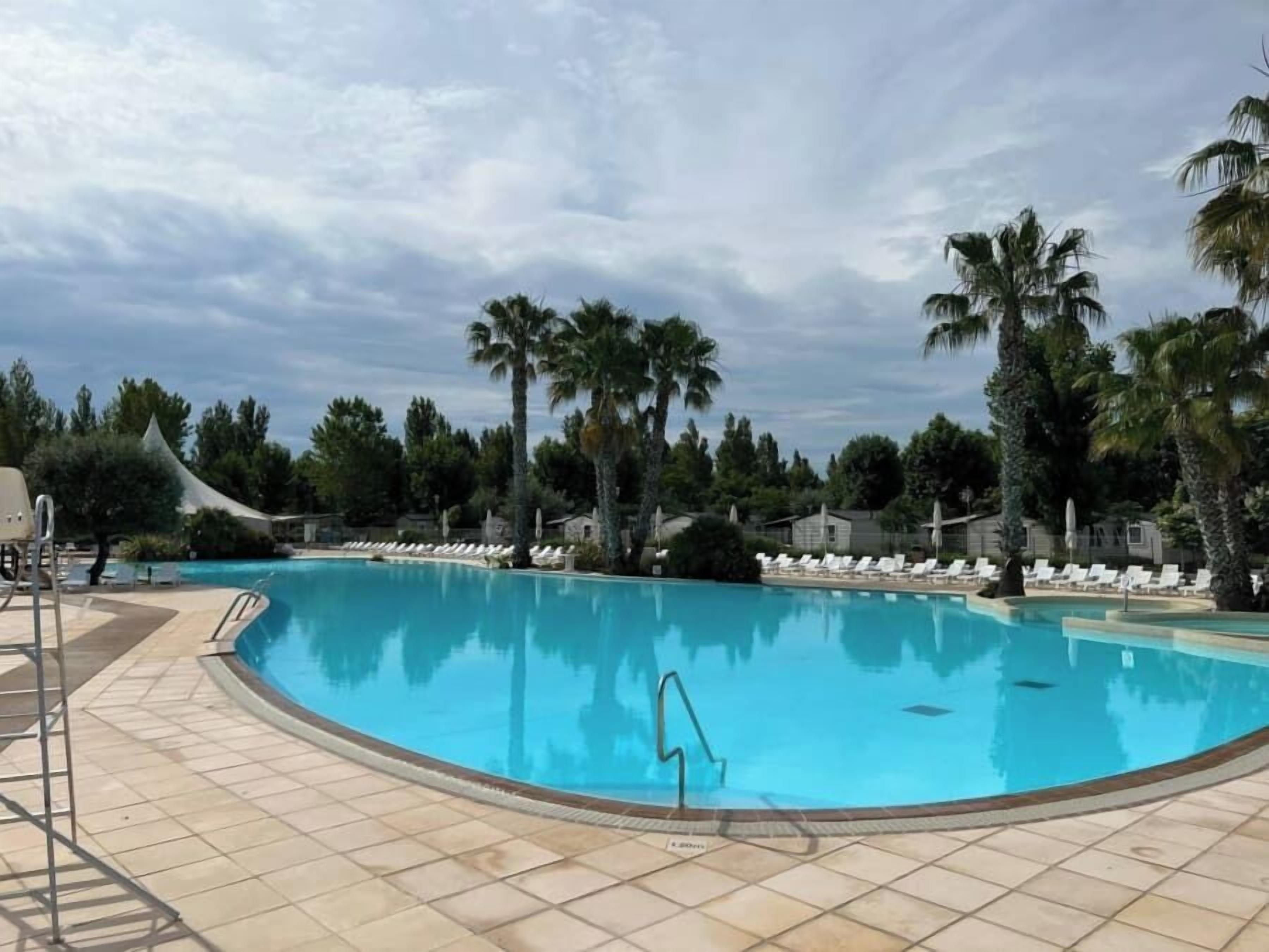 A large, clear swimming pool with a tiled deck, surrounded by palm trees and lounge chairs.