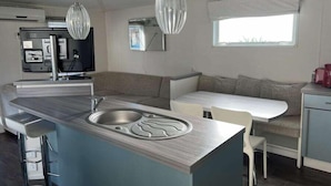 A modern kitchen with a central island, bar stools, a dining table, and pendant lighting. - Annick Mobilhome (Vias)
