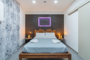 Une chambre d’hôtel moderne avec un grand lit, une tête de lit en bois, deux tables de chevet avec des lampes, un néon violet et un rideau à motifs floraux.