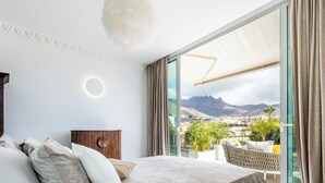 A bedroom with a large bed, a nightstand, and a view of a mountainous landscape through large windows. - New and Luxurious Villa Los Olivos  (Adeje)