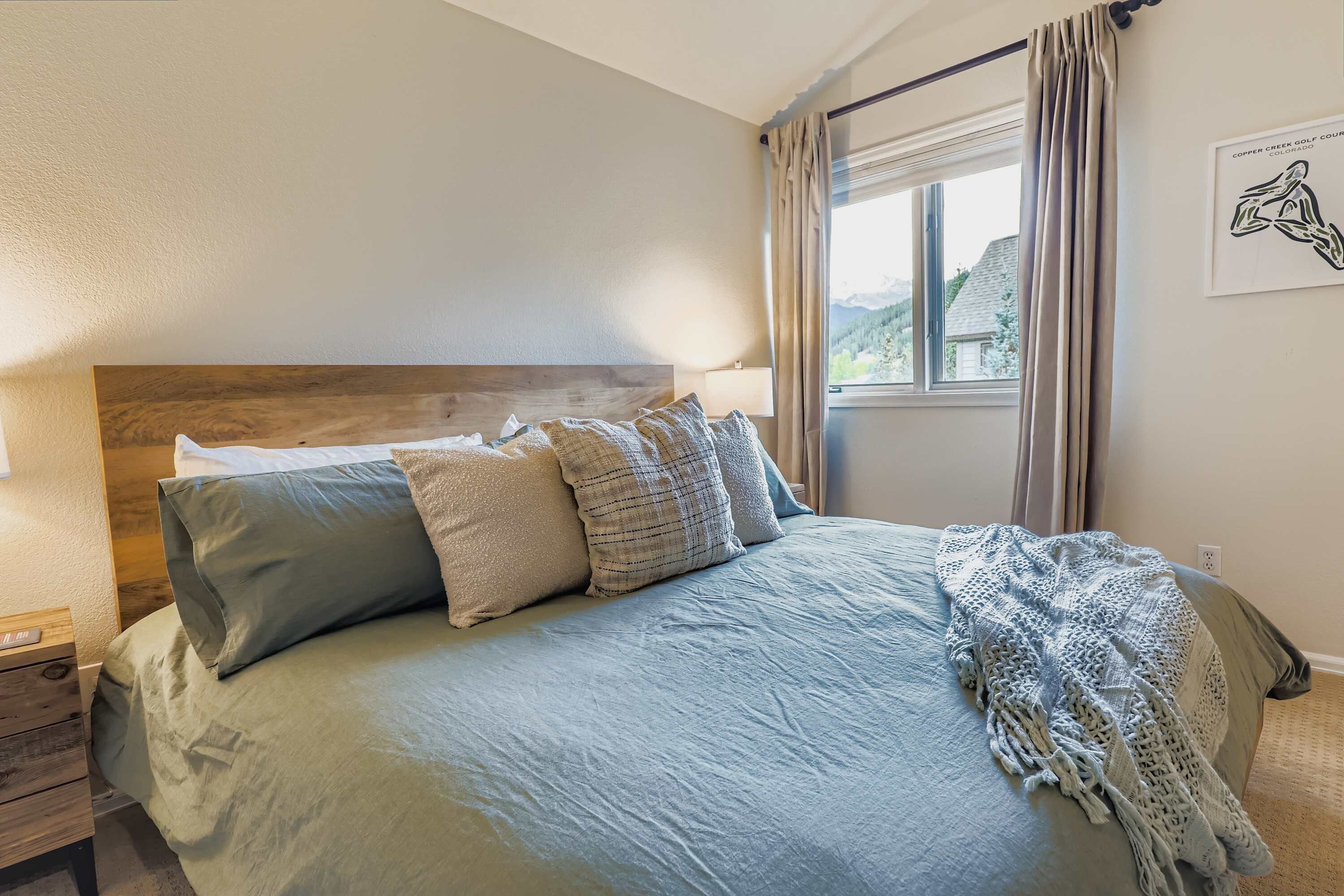 A bedroom with a wooden headboard, a bed with multiple pillows, a bedside table, a lamp, and a window with curtains.