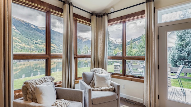 Habitación con amplios ventanales que ofrecen vistas a un lago y montañas. Hay dos sillones y un sofá con cojines.