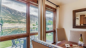 Habitación con ventanas amplias que ofrecen vistas a un lago y montañas. Hay una mesa con una botella de vino y copas, además de una silla con respaldo acolchado.