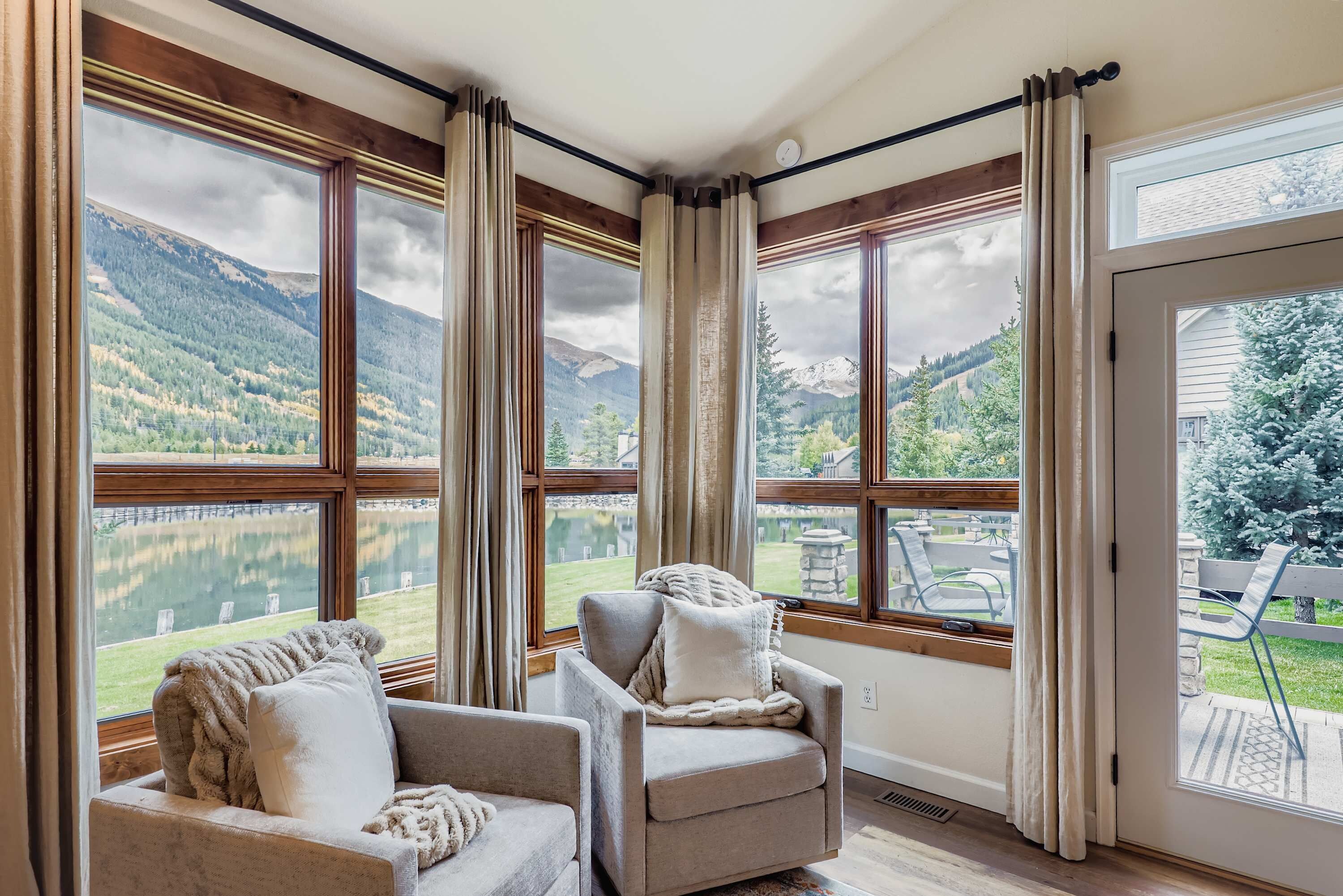 A room with large windows offering a view of a lake and mountains. There are two armchairs and a sofa with cushions.