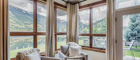 A room with large windows offering a view of a lake and mountains. There are two armchairs and a sofa with cushions.