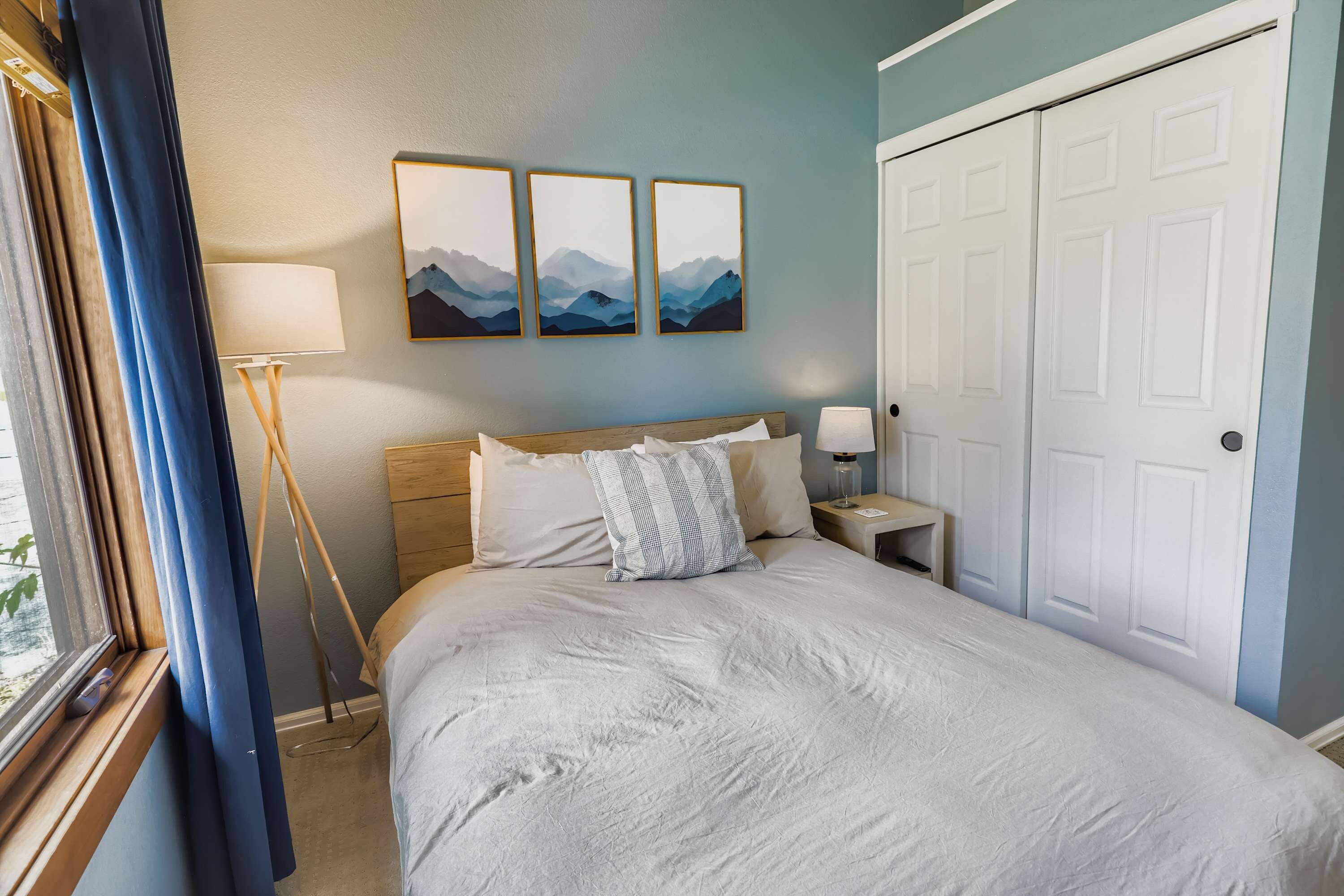 A neatly made bed with a white comforter and pillows, a bedside table with a lamp, and a wall with three framed paintings.