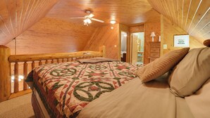 Un dormitorio acogedor en el ático con techo de madera, cama con colcha, ventilador de techo y vistas al pasillo.