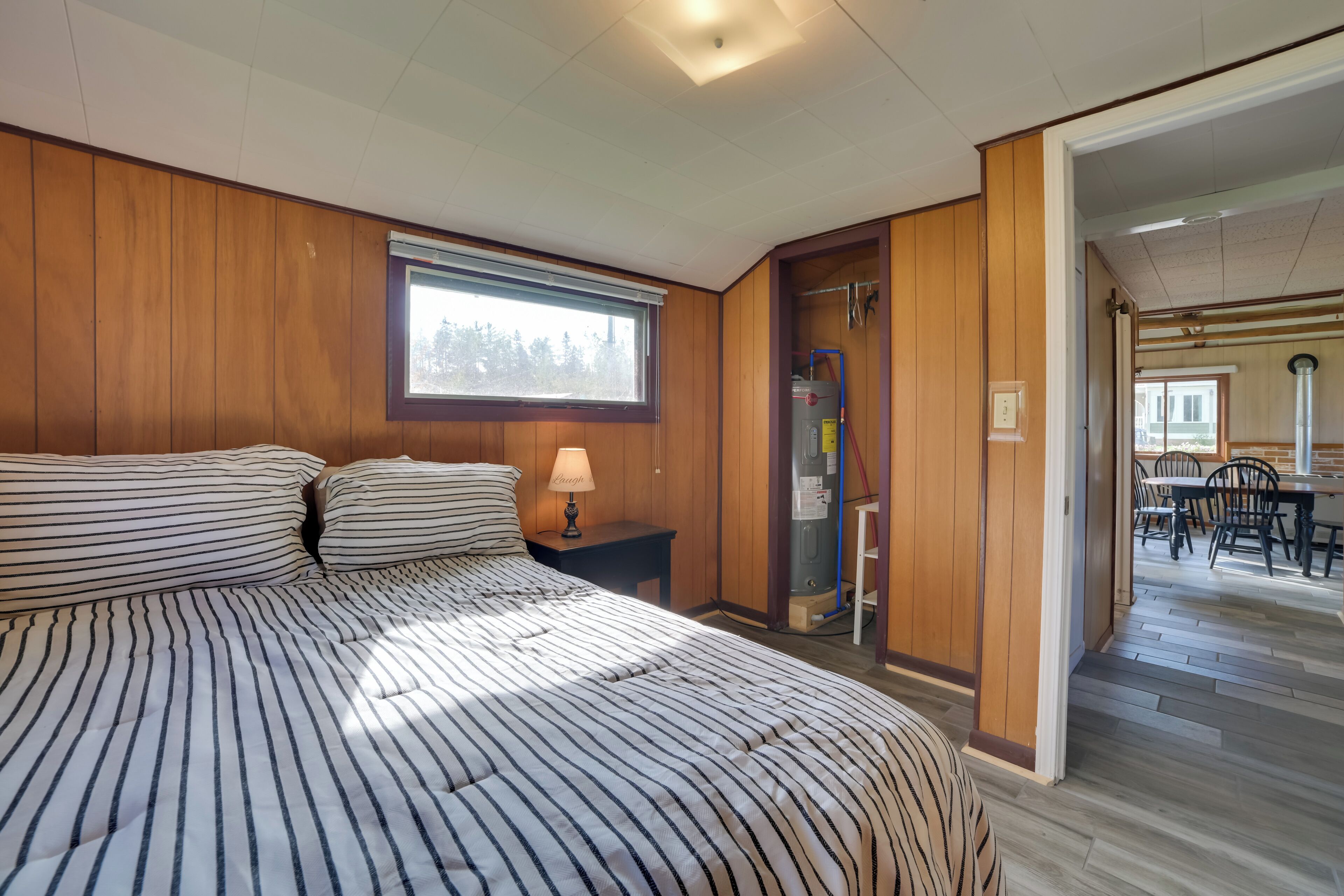 Une chambre avec un grand lit, une table de chevet, une lampe et une fenêtre. Une porte donne sur un coin repas équipé de chaises et d’une table.