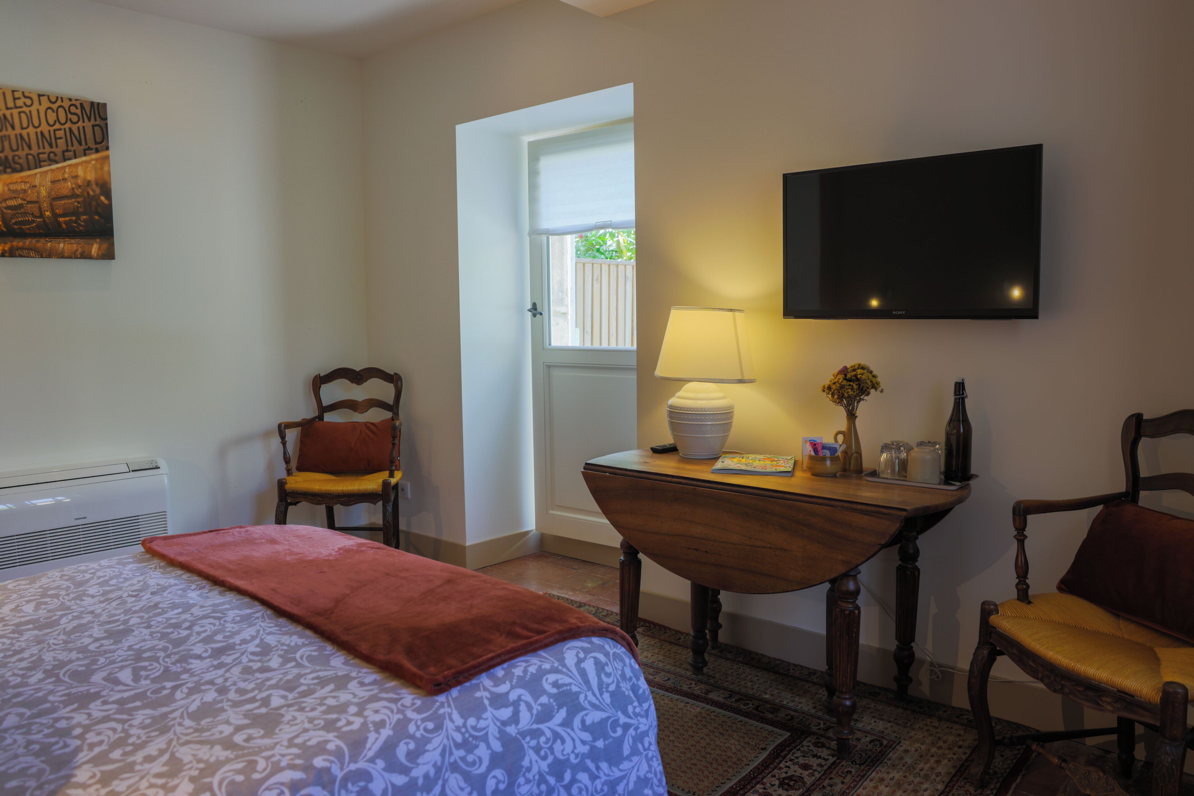 Habitación de hotel con cama, dos sillas, una mesa con lámpara, televisión y ventana con vista a zonas verdes.