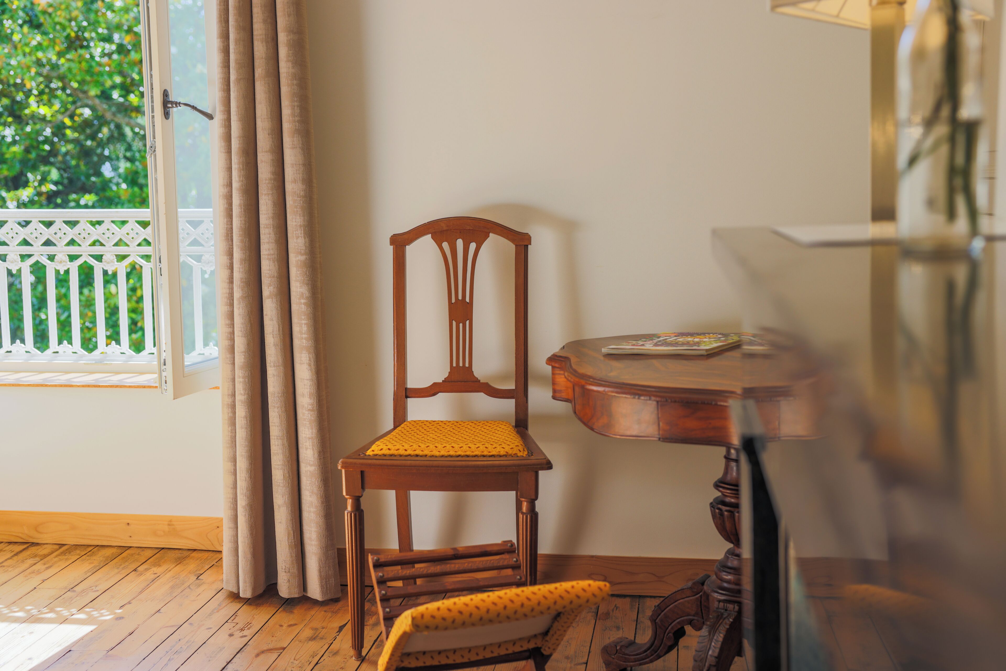 A room with a wooden chair with a yellow cushion, a small table with a book, and a window with curtains.