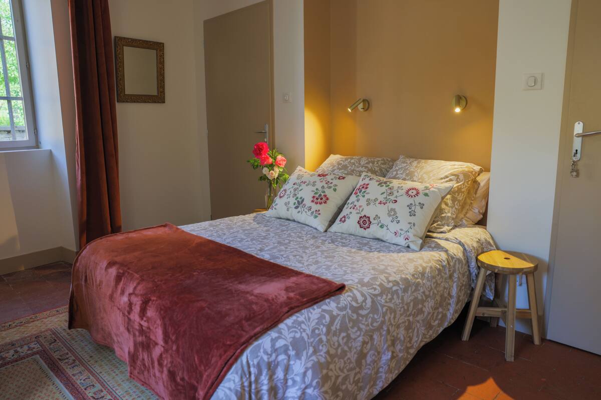 A bedroom with a bed, a bedside table, a mirror, and a flower vase.