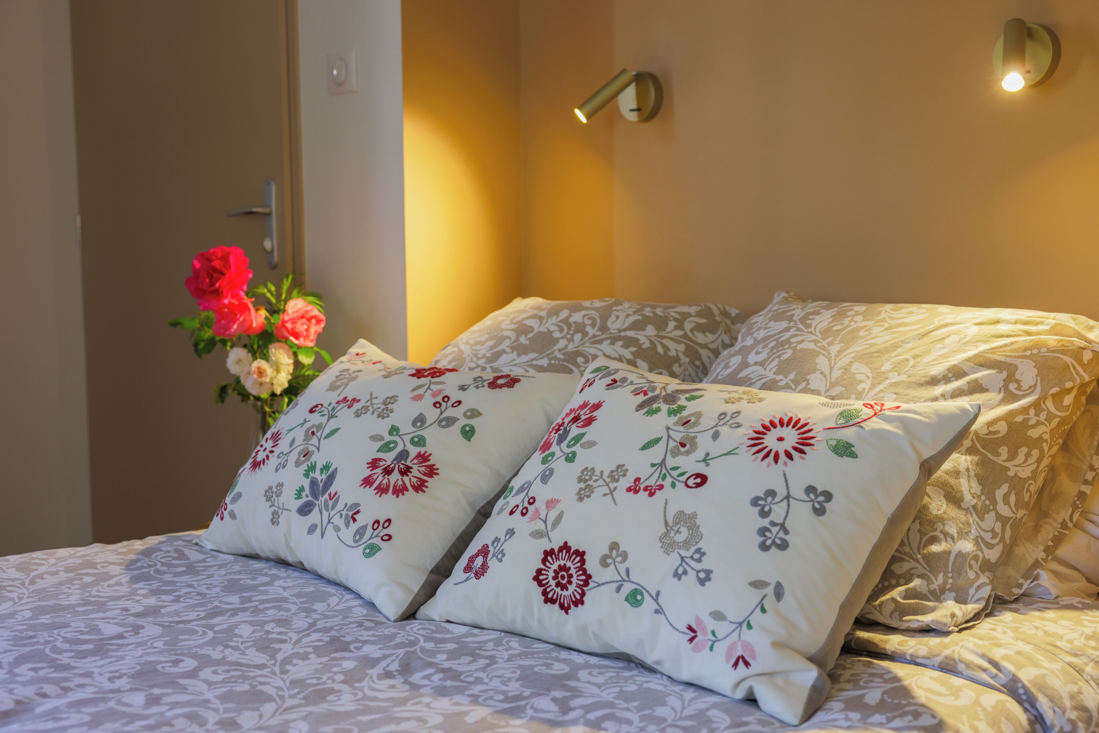 A neatly made bed with floral-patterned pillows and a vase of flowers on the side.