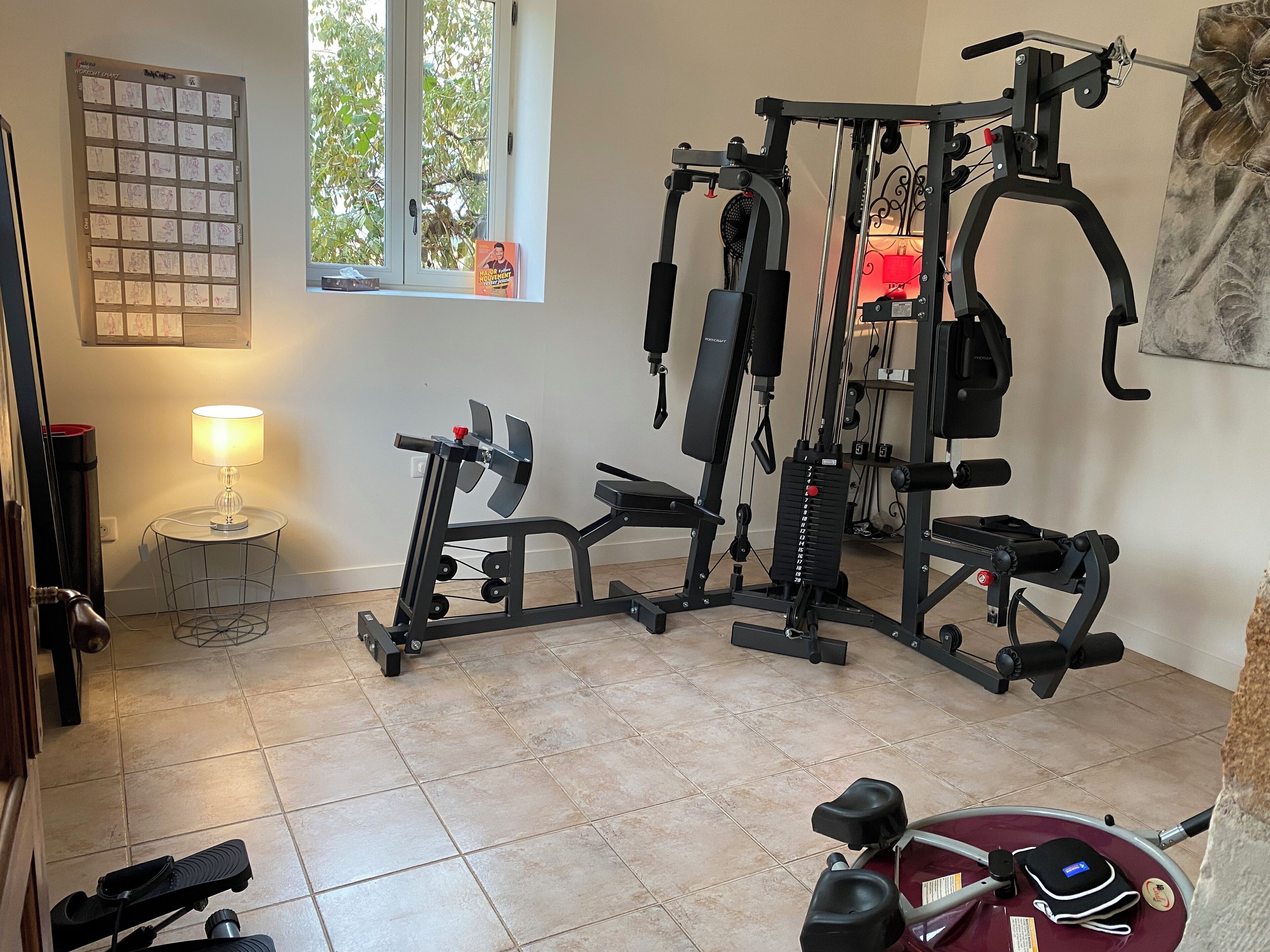 Een sportruimte met een multifunctionele fitnessapparaat, een hometrainer en een kleine tafel met een lamp.